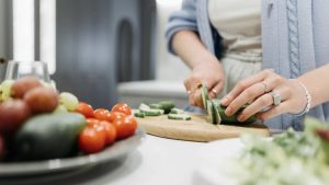 Close-up of a woman slicing cucumbers on a wooden board surrounded by fresh vegetables.